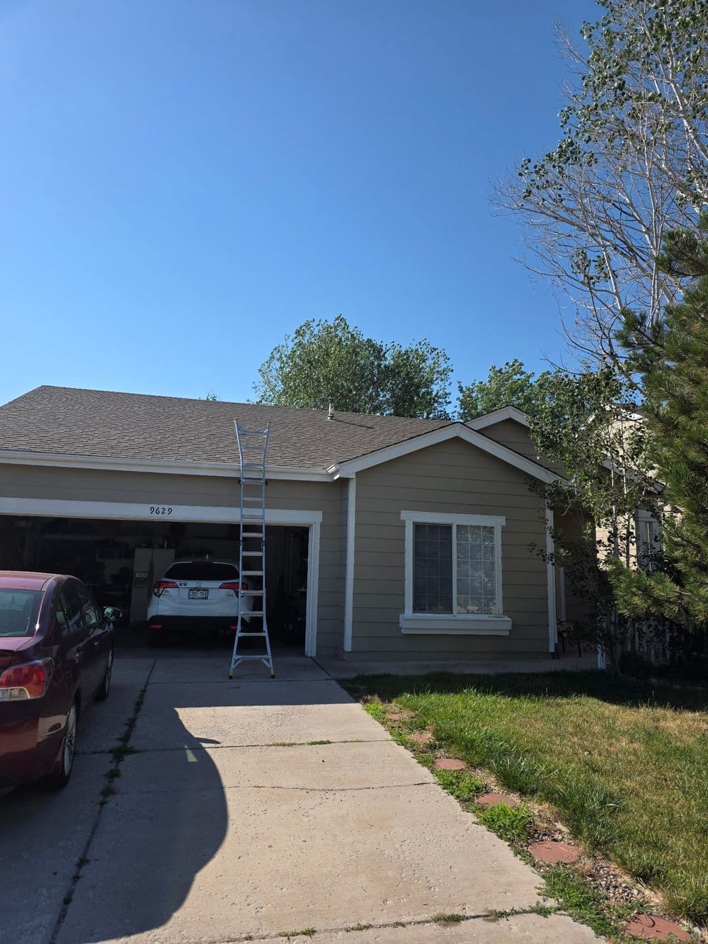 House with gray siding, parked car, ladder against the roof under a clear blue sky.